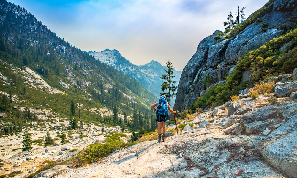 man in blue shirt and black shorts with hiking backpack walking on rocky mountain during daytime
