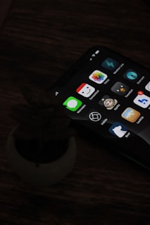 A smartphone with a dark screen displaying various app icons, including messages, photos, and other utilities is placed on a wooden table next to a small, indistinct object resembling a plant pot.