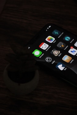 A smartphone with a dark screen displaying various app icons, including messages, photos, and other utilities is placed on a wooden table next to a small, indistinct object resembling a plant pot.