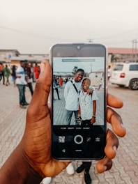 A hand holds a smartphone capturing an image of two people smiling, standing close together outdoors. The background shows several people walking and parked cars on a paved area, suggesting a social gathering.