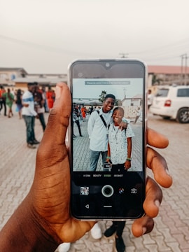 A hand holds a smartphone capturing an image of two people smiling, standing close together outdoors. The background shows several people walking and parked cars on a paved area, suggesting a social gathering.