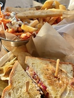 A close-up of a hearty meal featuring a grilled sandwich with a crispy golden crust, filled with melted cheese and toppings, secured with a skewer. Alongside are a variety of fries including regular and sweet potato, garnished with blue cheese crumbles. The setting includes brown parchment paper beneath the food.