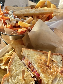 A close-up of a hearty meal featuring a grilled sandwich with a crispy golden crust, filled with melted cheese and toppings, secured with a skewer. Alongside are a variety of fries including regular and sweet potato, garnished with blue cheese crumbles. The setting includes brown parchment paper beneath the food.