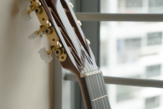A close-up view of a classical guitar's headstock, showing the tuning pegs in detail. The guitar is leaning against a wall with a window in the background, partially out of focus.