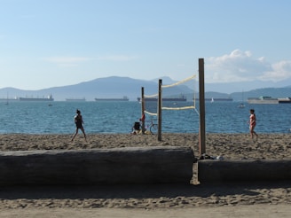 A sunny beach scene with people engaging in recreational activities. Two individuals are near a volleyball net, with a bicycle parked nearby. In the background, several large ships are visible on the water, with mountains and blue sky creating a serene backdrop.