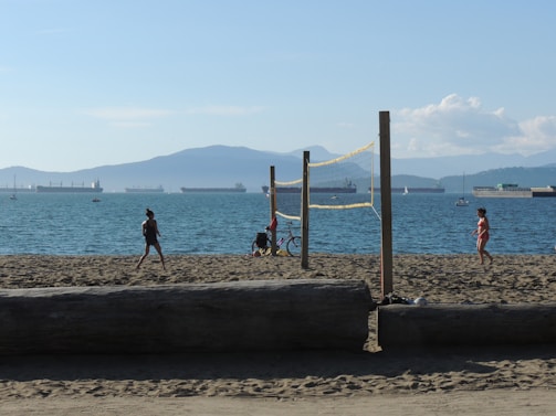 A sunny beach scene with people engaging in recreational activities. Two individuals are near a volleyball net, with a bicycle parked nearby. In the background, several large ships are visible on the water, with mountains and blue sky creating a serene backdrop.