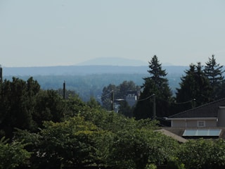 A landscape with a residential area partially visible in the foreground, featuring rooftops and lush trees. In the distance, layers of hills and a faint outline of a mountain are visible under a clear sky. The scene is serene and showcases a natural and suburban blend.