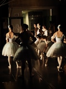 A group of ballerinas backstage, preparing for a performance. They are dressed in white tutus with blue accents and have their hair neatly styled. Some are standing while others are adjusting their attire, creating a scene of anticipation and focus.