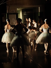 A group of ballerinas backstage, preparing for a performance. They are dressed in white tutus with blue accents and have their hair neatly styled. Some are standing while others are adjusting their attire, creating a scene of anticipation and focus.