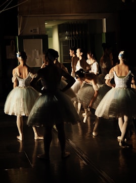 A group of ballerinas backstage, preparing for a performance. They are dressed in white tutus with blue accents and have their hair neatly styled. Some are standing while others are adjusting their attire, creating a scene of anticipation and focus.