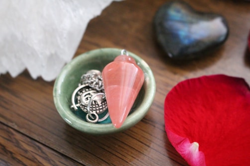A small green ceramic bowl holds a pink stone pendulum and some metallic jewelry, including a chain and a round pendant. Next to the bowl, there is a red rose petal and a dark, heart-shaped stone with an iridescent surface. An edge of a larger white crystal is visible in the background, and the items are placed on a wooden surface.