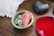 A small green ceramic bowl holds a pink stone pendulum and some metallic jewelry, including a chain and a round pendant. Next to the bowl, there is a red rose petal and a dark, heart-shaped stone with an iridescent surface. An edge of a larger white crystal is visible in the background, and the items are placed on a wooden surface.