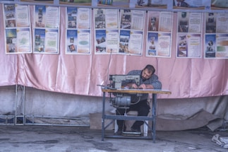 A man is seated at a sewing machine, focused on his work, against a backdrop of informational posters. The environment appears to be a workshop or an outdoor stall with a fabric backdrop, possibly in a community setting.