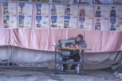 A man is seated at a sewing machine, focused on his work, against a backdrop of informational posters. The environment appears to be a workshop or an outdoor stall with a fabric backdrop, possibly in a community setting.
