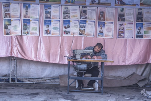 A man is seated at a sewing machine, focused on his work, against a backdrop of informational posters. The environment appears to be a workshop or an outdoor stall with a fabric backdrop, possibly in a community setting.