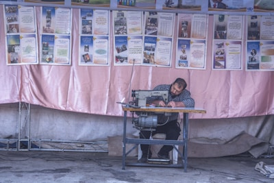 A man is seated at a sewing machine, focused on his work, against a backdrop of informational posters. The environment appears to be a workshop or an outdoor stall with a fabric backdrop, possibly in a community setting.