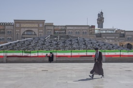 Two individuals are present in an urban setting, with one sitting near a railing adorned with the Iranian flag and the other walking in the foreground. Behind them is a large, modern glass structure with an intricate design, and traditional architectural buildings are visible in the background.