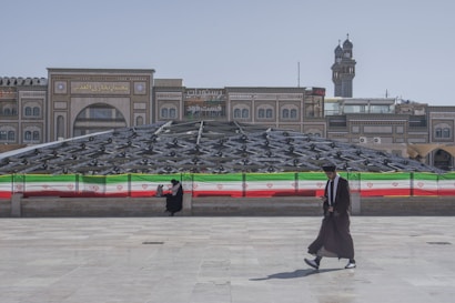 Two individuals are present in an urban setting, with one sitting near a railing adorned with the Iranian flag and the other walking in the foreground. Behind them is a large, modern glass structure with an intricate design, and traditional architectural buildings are visible in the background.
