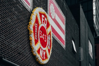A large floral display shaped like a firefighter badge with 'FDNY 343' in the center is affixed to a dark brick wall. The display is bordered with yellow flowers and predominantly features red and white flowers. Above it, a red and white striped banner is visible with a logo. The scene is urban and somber.