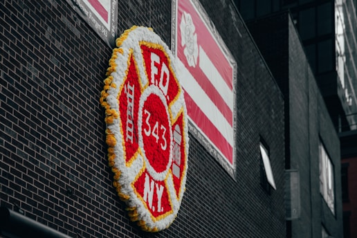 A large floral display shaped like a firefighter badge with 'FDNY 343' in the center is affixed to a dark brick wall. The display is bordered with yellow flowers and predominantly features red and white flowers. Above it, a red and white striped banner is visible with a logo. The scene is urban and somber.