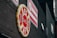 A large floral display shaped like a firefighter badge with 'FDNY 343' in the center is affixed to a dark brick wall. The display is bordered with yellow flowers and predominantly features red and white flowers. Above it, a red and white striped banner is visible with a logo. The scene is urban and somber.