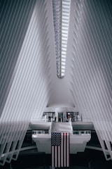 A futuristic architectural interior with a symmetrical design features white, ribbed walls converging towards the ceiling. The prominent structure is bright with natural light streaming in through a linear skylight. A large American flag hangs from a central platform, with a few people walking around near the entrance or crossing a suspended bridge-like walkway.