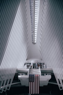 A futuristic architectural interior with a symmetrical design features white, ribbed walls converging towards the ceiling. The prominent structure is bright with natural light streaming in through a linear skylight. A large American flag hangs from a central platform, with a few people walking around near the entrance or crossing a suspended bridge-like walkway.