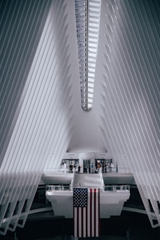 A futuristic architectural interior with a symmetrical design features white, ribbed walls converging towards the ceiling. The prominent structure is bright with natural light streaming in through a linear skylight. A large American flag hangs from a central platform, with a few people walking around near the entrance or crossing a suspended bridge-like walkway.