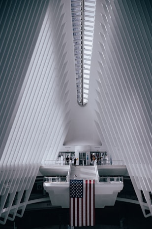 A futuristic architectural interior with a symmetrical design features white, ribbed walls converging towards the ceiling. The prominent structure is bright with natural light streaming in through a linear skylight. A large American flag hangs from a central platform, with a few people walking around near the entrance or crossing a suspended bridge-like walkway.
