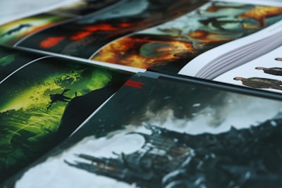A collection of art prints displayed in layers, with vibrant colors and dynamic scenes. The foreground features a fantasy landscape in green hues with a silhouette of a creature. Other sections display fiery imagery and detailed character designs.