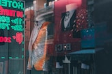 A stylish storefront display featuring two mannequins wearing ornate suits. The suit on the left is predominantly silver with orange accents, embellished with intricate patterns. The suit on the right is a black jacket with colorful sparkling details. A reflections of neon signs with eyecatching text is visible in the glass.
