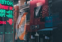 A stylish storefront display featuring two mannequins wearing ornate suits. The suit on the left is predominantly silver with orange accents, embellished with intricate patterns. The suit on the right is a black jacket with colorful sparkling details. A reflections of neon signs with eyecatching text is visible in the glass.