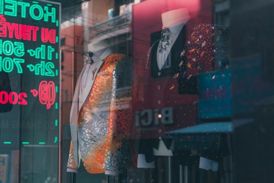 A stylish storefront display featuring two mannequins wearing ornate suits. The suit on the left is predominantly silver with orange accents, embellished with intricate patterns. The suit on the right is a black jacket with colorful sparkling details. A reflections of neon signs with eyecatching text is visible in the glass.