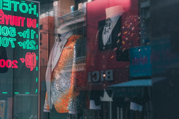 A stylish storefront display featuring two mannequins wearing ornate suits. The suit on the left is predominantly silver with orange accents, embellished with intricate patterns. The suit on the right is a black jacket with colorful sparkling details. A reflections of neon signs with eyecatching text is visible in the glass.