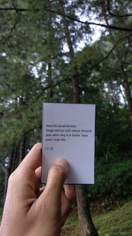 A hand is holding a card with text in a natural setting. Tall trees with lush green foliage fill the background, suggesting a forest environment. The card contains a message in a non-English language.