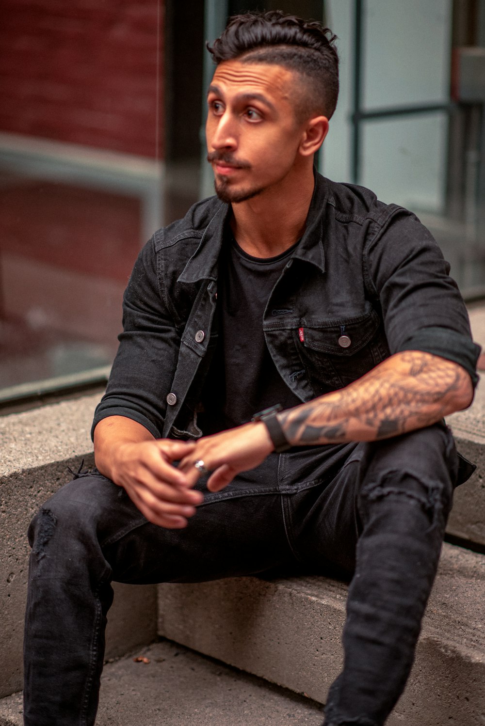 man in black leather jacket and black denim jeans sitting on concrete floor