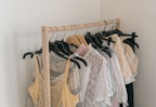 A wooden clothing rack holds an assortment of light, airy garments. The clothes include a variety of dresses and tops in pastel colors, featuring lace and delicate fabrics. Some hangers are wooden while others are black plastic.