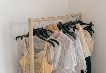 A wooden clothing rack holds an assortment of light, airy garments. The clothes include a variety of dresses and tops in pastel colors, featuring lace and delicate fabrics. Some hangers are wooden while others are black plastic.