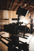 A professional-grade video camera mounted on a tripod is set up in an indoor environment with a wood-paneled ceiling. The background is softly blurred, revealing a person in casual attire, possibly adjusting equipment or working in the setting.