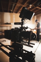 A professional-grade video camera mounted on a tripod is set up in an indoor environment with a wood-paneled ceiling. The background is softly blurred, revealing a person in casual attire, possibly adjusting equipment or working in the setting.