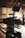 A professional-grade video camera mounted on a tripod is set up in an indoor environment with a wood-paneled ceiling. The background is softly blurred, revealing a person in casual attire, possibly adjusting equipment or working in the setting.