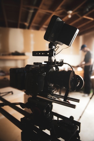 A professional-grade video camera mounted on a tripod is set up in an indoor environment with a wood-paneled ceiling. The background is softly blurred, revealing a person in casual attire, possibly adjusting equipment or working in the setting.