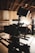 A professional-grade video camera mounted on a tripod is set up in an indoor environment with a wood-paneled ceiling. The background is softly blurred, revealing a person in casual attire, possibly adjusting equipment or working in the setting.