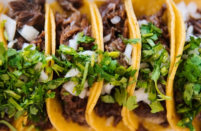 Four tacos are filled with shredded meat, garnished with chopped onions and fresh cilantro, all wrapped in soft corn tortillas.
