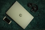 A closed HP laptop is placed on a textured green surface, alongside a black smartphone and a pair of headphones. The elements are arranged with the laptop centrally positioned, the phone to the bottom left, and the headphones to the top right. The color contrast between the light silver laptop and the dark accessories creates a balanced composition.