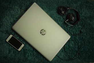 A closed HP laptop is placed on a textured green surface, alongside a black smartphone and a pair of headphones. The elements are arranged with the laptop centrally positioned, the phone to the bottom left, and the headphones to the top right. The color contrast between the light silver laptop and the dark accessories creates a balanced composition.