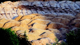 A rugged landscape of eroded hills and ridges, with layers of light brown and darker brown soil visible. The texture suggests a natural terrain, possibly a quarry or a desert-like area. In the foreground, green foliage adds contrast to the earthy tones.