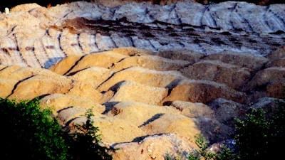 A rugged landscape of eroded hills and ridges, with layers of light brown and darker brown soil visible. The texture suggests a natural terrain, possibly a quarry or a desert-like area. In the foreground, green foliage adds contrast to the earthy tones.