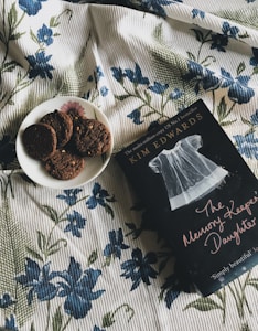 A book titled 'The Memory Keeper's Daughter' lies on a floral-patterned blanket next to a plate with several chocolate cookies. The fabric of the blanket is adorned with blue flowers and green leaves.