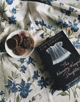 A book titled 'The Memory Keeper's Daughter' lies on a floral-patterned blanket next to a plate with several chocolate cookies. The fabric of the blanket is adorned with blue flowers and green leaves.